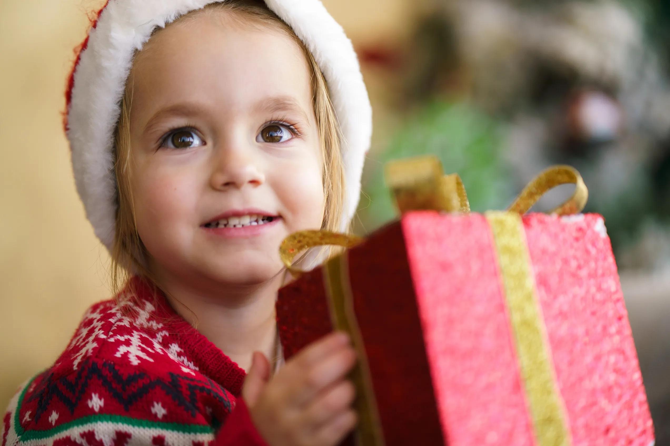 Kind im Weihnachtspullover und mit Weihnachtsmütze hält Geschenk in den Händen.